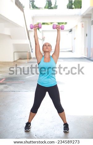 Back view of sporty woman holding pink barbell with both arms stretched out for shoulder strengthening, outdoors.