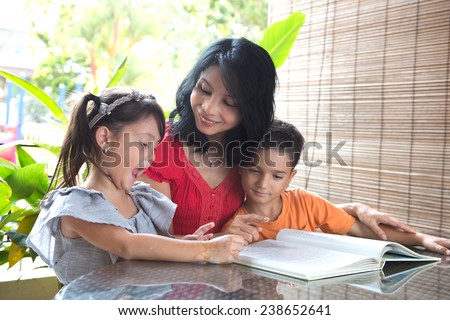 Asian mother with young daughter and son reading a story book together in a home environment