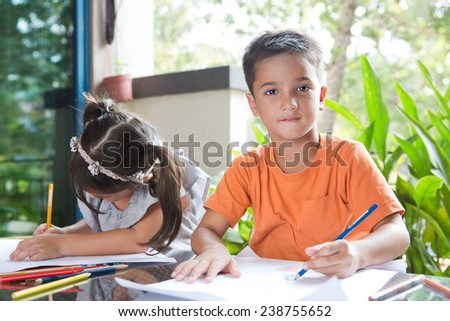 Cute little pan asian boy smiling while holding a color pencil sitting next to his younger sister engross in her coloring activity in home environment