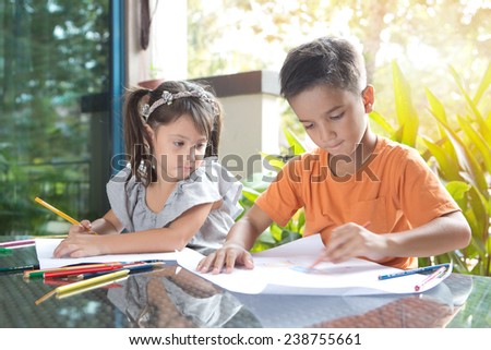 Cute little pan asian boy coloring his drawing while sitting while being watched by his curious younger sister in home environment