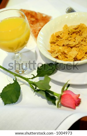 A tray of breakfast consisting of a glass of orange juice , cornflakes, a round of toast and a stalk of pink rose