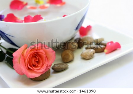 A setting for a sensual body massage consisting of a bowl of sensual floral oil mix with flower petals, pebbles and candle