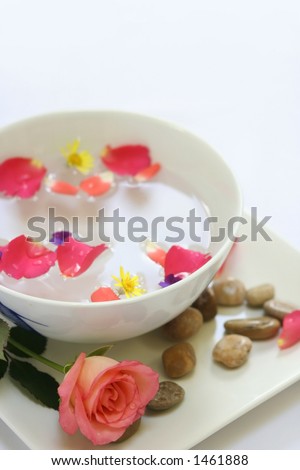 A setting for a sensual body massage consisting of a bowl of sensual floral oil mix with flower petals, pebbles and candle
