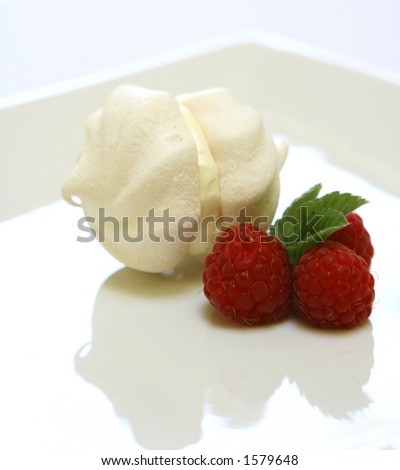 A double cup of meringue filled with cream on a plate with three raspberries as decoration