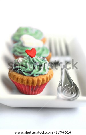 Two mini cupcakes with butter icing and silver ball sprinkles, served on white plate and a metal fork, isolated on white