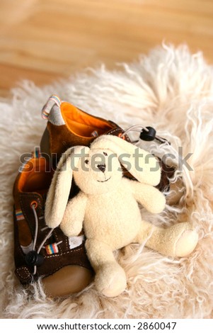 A fluffy toy bunny resting on a pair of toddler\'s shoes on sheepskin rug.