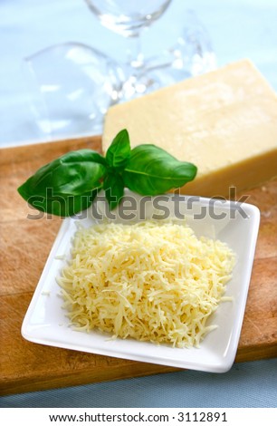 A bowl of grated mature cheddar cheese on wooden chopping board with block of cheese in background and spring of fresh green basil.