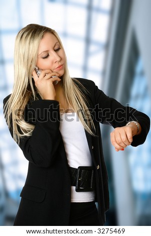 Young businesswoman looking at her watch while talking on her mobile phone aginst interior corporate background. Concept of business and time pressure.