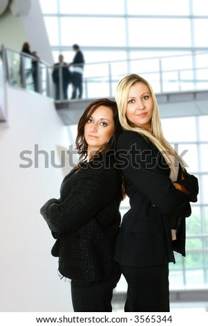 Two caucasian businesswoman standing back to back against interior corporate building. Concept of teamwork and profesionalism.