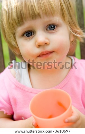 Curious girl toddler sitting outdoors, with curious eyes, looking at viewer, holding an orange plastic cup.