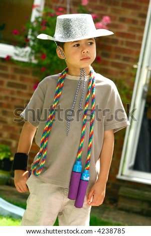Young boy having fun dressing up, with a sparkly cowboy hat and skipping rope as pretend lasso, outdoor setting.