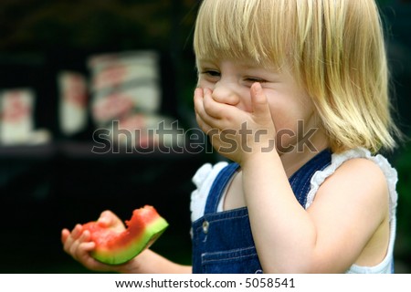 Laughing toddler girl with one hand covering mouth while another holding a piece of juicy red watermelon, having fun in the garden.