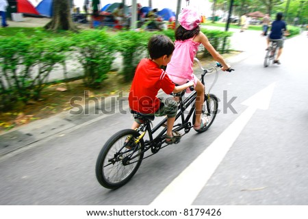 Two young children riding on a tandem bike in a public park