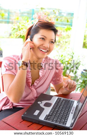 Young Asian businesswoman talking on her cellphone with laptop on table, in an outdoor environment