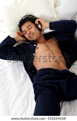 Handsome young nepalese man in pajamas, laid-back  in bed listening music in headphones . Studio shot. White background.