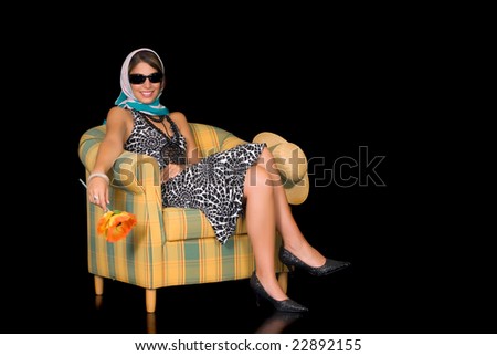Attractive young classy retro style woman, lady sitting in arm chair holding flower.  Studio shot, black background.