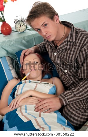 Young attractive woman sick in  bed, thermometer in mouth. Man nursing wife. Studio.