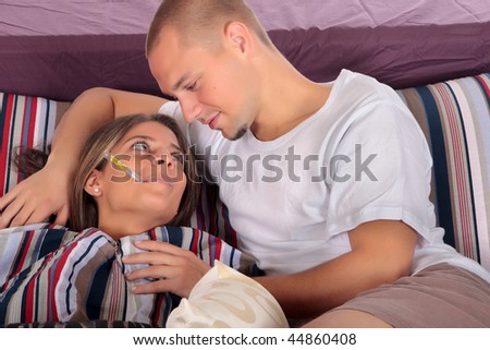 Young attractive woman sick in  bed, thermometer in mouth. Man nursing wife. Studio.