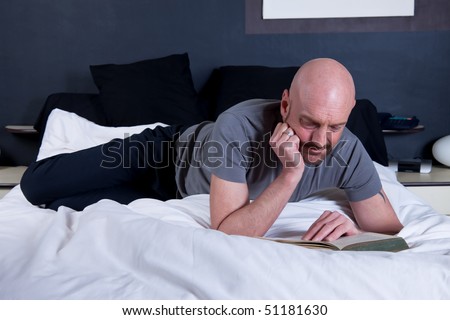 Bald Middle aged man reading book on bed in bedroom.