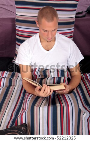 Young handsome man enjoying and reading a book  in bed in bedroom.  Studio.