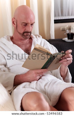 Bald Middle aged man in bathrobe at home relaxing in sofa reading a book, artistic composition, shallow dof, focus on man.