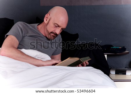 Bald Middle aged man reading book on bed in bedroom.
