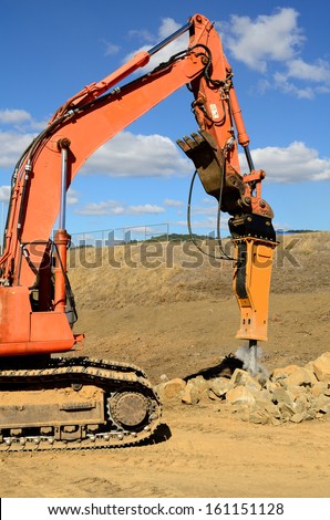 A large hydraulic rock breaker ram hammer installed on a track hoe excavator being used to break up stones at a new commercial construction development project