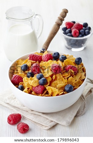 Bowl of corn flakes and fresh berries and jug of milk on white wooden background