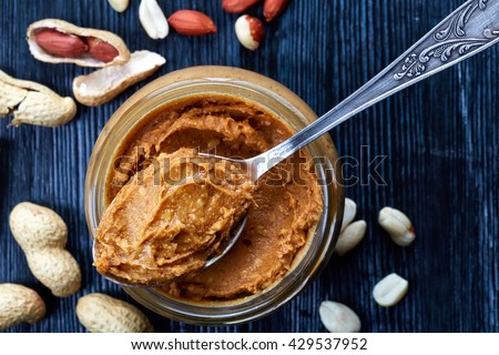 Jar and spoon of peanut butter and peanuts on dark wooden background from top view