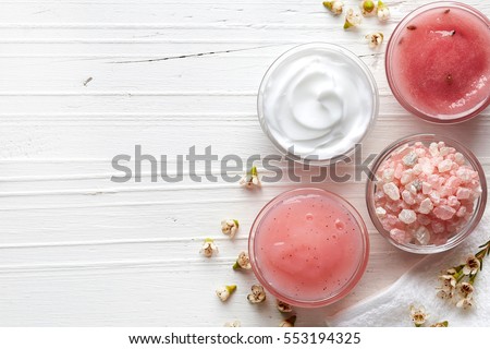 Cosmetic cream container, body scrub, bath salt and pink flowers on white wooden background from top view