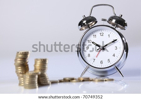 Time is money concept. Gold pound coins in foreground blurred, with a traditional silver and chrome alarm clock in background in sharp focus.