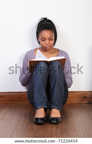 Beautiful young black woman wearing jeans and purple top, sitting on the floor at home, reading a book. She is wearing a purple, long sleeved top and blue denim jeans.