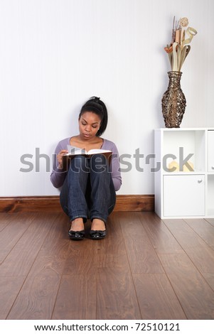 Beautiful young black woman wearing jeans and purple top, sitting on the floor at home, reading a book.