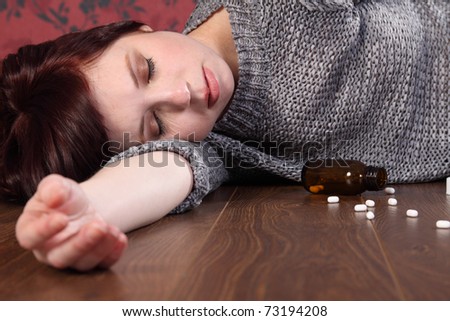 Teenager girl lying on the floor at home after an overdose of pills. Her eyes are closed and there is a bottle of pills on the floor beside her.