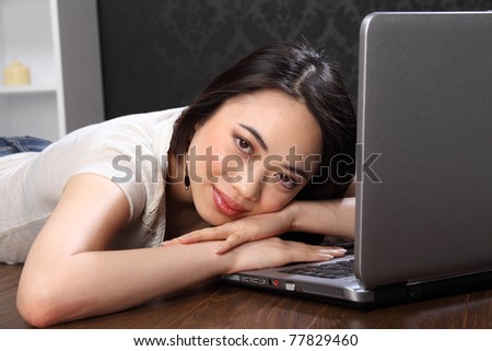 Resting on the keyboard, a beautiful smiling young chinese asian girl lying on the floor at home, surfing the internet on her laptop computer.