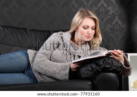 Quiet time for beautiful young blonde woman sitting reading a book on black leather sofa at home, wearing casual grey knitted sweater, blue jeans and just relaxing.
