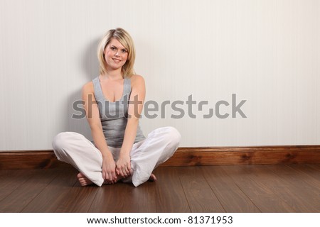 Beautiful young caucasian woman with blonde hair and happy smile sitting cross legged on laminate floor at home, relaxing bare foot. She is wearing white linen pants and a grey vest.