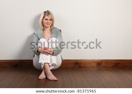 Beautiful young caucasian woman with blonde hair and happy smile sitting on laminate floor at home, relaxing bare foot. She is wearing white linen pants and a grey heavy knit cardigan.