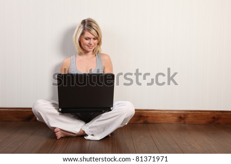 Sitting cross legged on laminate floor at home a beautiful young blonde caucasian girl has fun surfing the internet on her laptop. She is bare foot in white linen trousers and grey vest.
