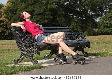 Sitting on park bench a beautiful young woman with long legs wearing roller skates relaxes with her eyes closed, enjoying the warm summer sunshine. She is wearing a red top and denim cut off shorts.