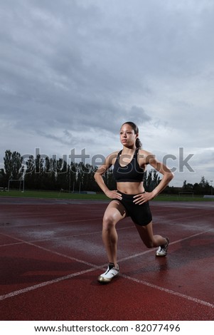 Warm up lunge exercise for quadriceps by fit young female athlete on athletics running track, wearing black lycra sports outfit and running spikes. Grey cloudy sky in background.