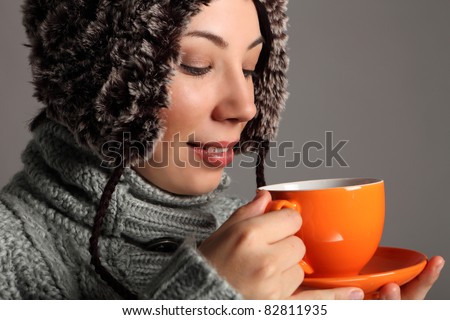 Beautiful young woman wearing thick wool sweater and furry winter hat, drinking tea from orange cup. Shot against a grey background.