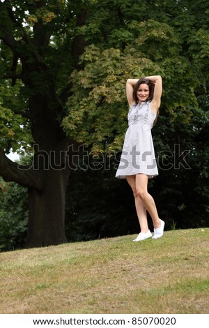 Happy moment enjoying nature for slim young woman wearing short dress outdoors in countryside with arms outstretched, enjoying sunshine near large forest trees.