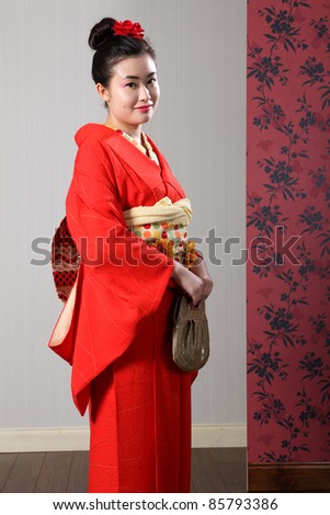 Kimono model beautiful young oriental woman wearing red Japanese traditional kimono, a full length robe garment complete with obi sash and fashion bag. Model wearing red kanzashi flower in hair.