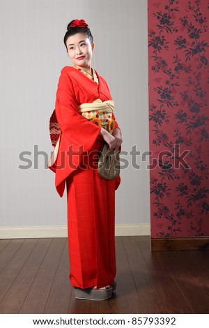 Traditional red Japanese kimono robe garment complete with obi sash being modeled by beautiful young asian oriental model with kanzashi hair flower. Shot indoors.
