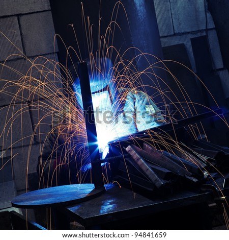 Worker wearing protective face mask using mig welding causing sparks flying over the working table while producing metal details