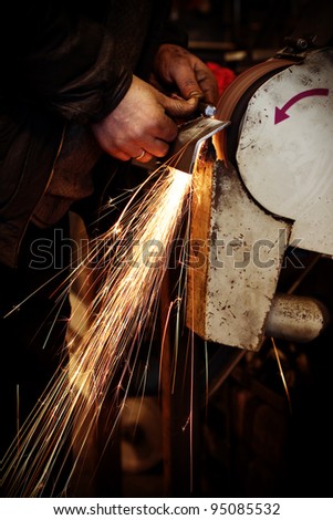 Worker in a factory keeping a metal plate in his hands and grinding it on grinder, causing many sparks