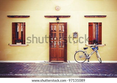 Pretty lonely bike by classic style house entrance (yellow walls, wooden door and windows decorated with flowerpots), vintage aged blurry photo effect added