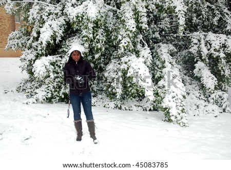 African girl standing in front of snowy bush making snow ball in winter and watching at camera