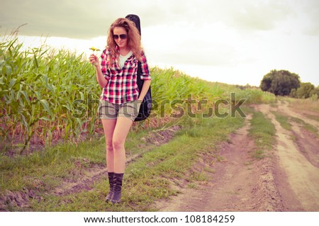 Young redhead woman with guitar passes corn field outdoors in summer by dirt road. Split toning.